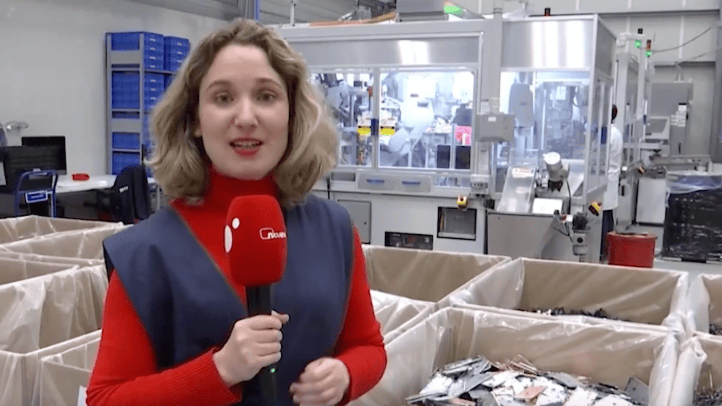 Woman reporter with microphone in factory setting, manufacturing machinery in background.